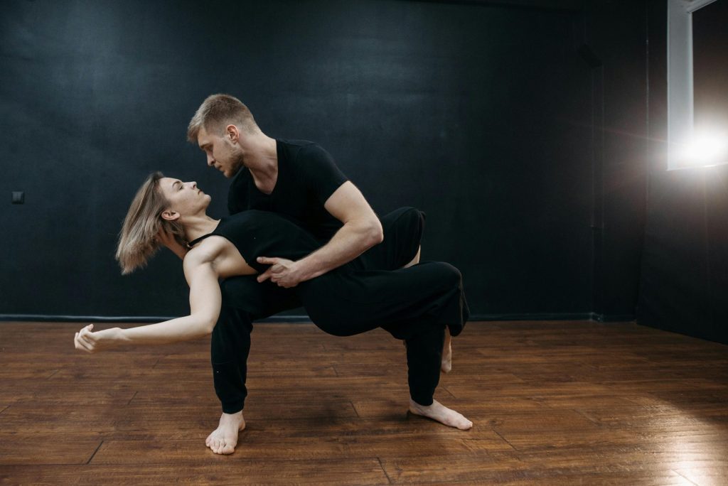 A couple in black attire exhibiting graceful dance moves on wooden flooring indoors.